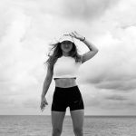 Woman in sportswear stands confidently by the sea against a cloudy sky.